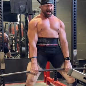 maxresdefault A woman in black gym wear performing a heavy barbell deadlift in a rustic, brick-walled garage gym. She has her hands chalked and is maintaining a neutral spine in the starting position of the lift.