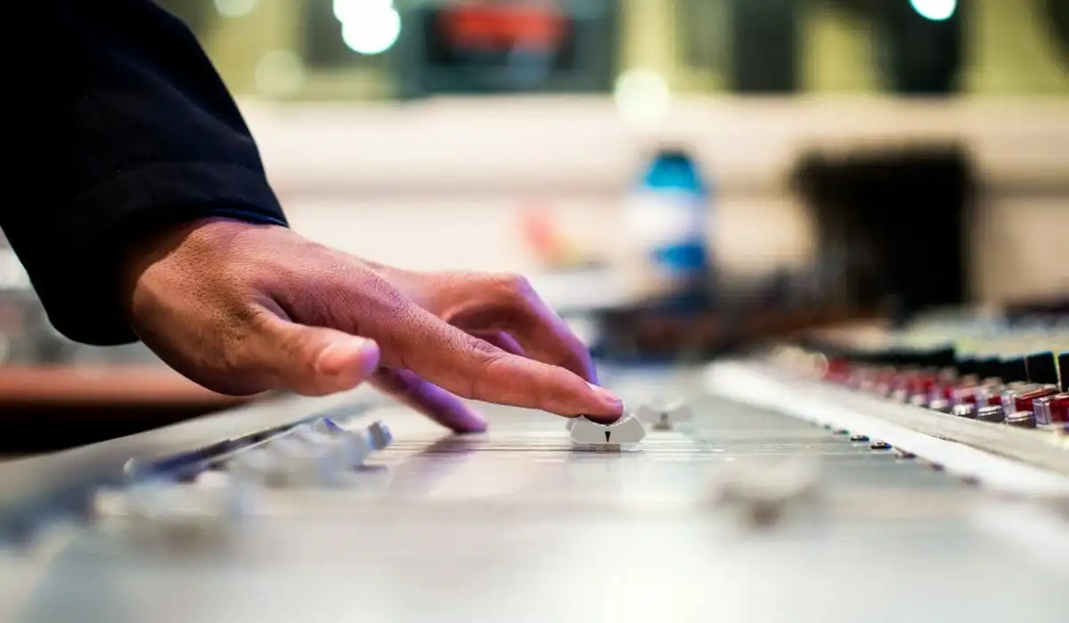 A close-up photograph of a hand adjust a grey fader on a large professional audio mixing console. The background is a brightly lit and blurred music recording studio.