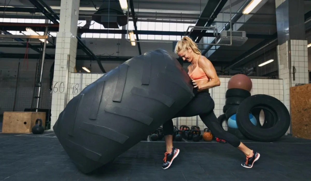 A strong woman in athletic gear performing a tire flip in a gym setting, demonstrating power and functional strength training.