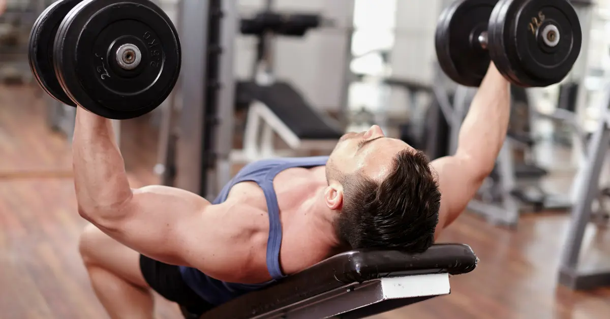 A man in a blue tank top performing an incline dumbbell bench press on a weight bench in a gym. He is shown mid-rep with arms extended, holding heavy black dumbbells.