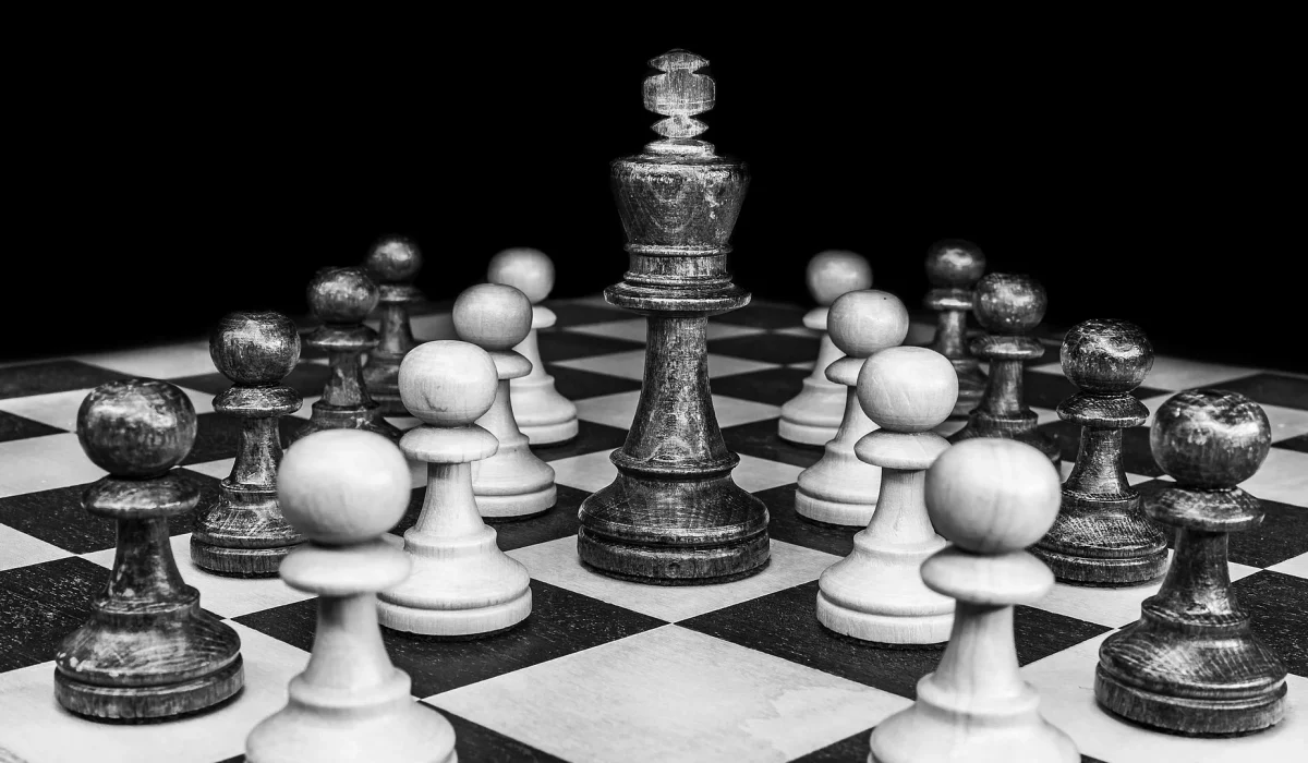 A monochromatic close-up of a chess board with a large black king piece standing at the center, surrounded by a circle of smaller white and black pawn pieces.