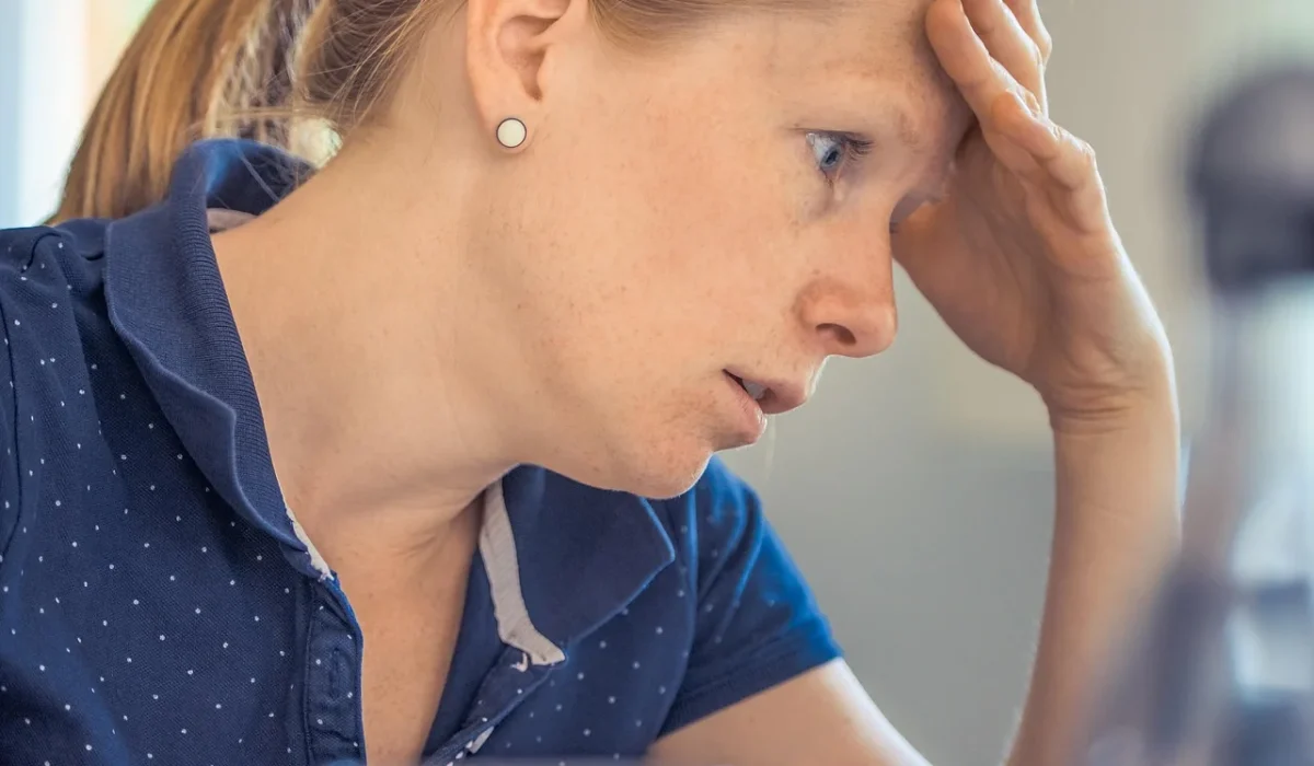 A side profile of a woman with her hair in a ponytail, sitting at a desk and leaning forward with her hand on her forehead while looking intently at a computer screen.