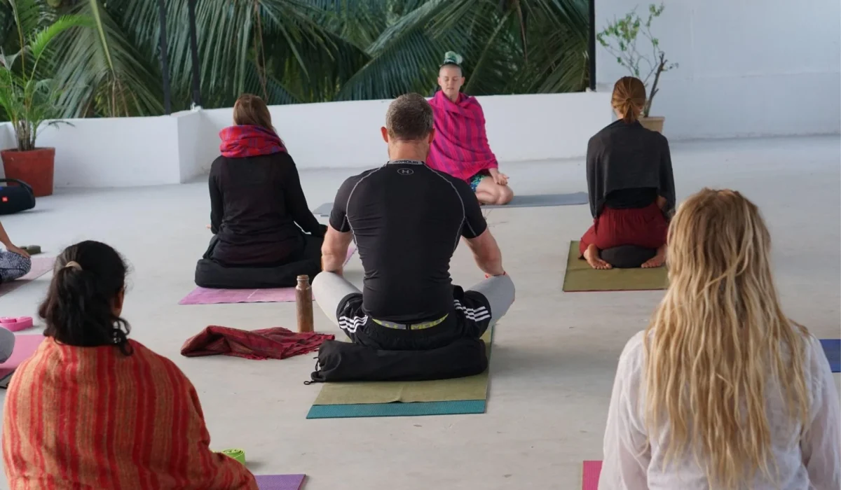 A view from behind a group of people sitting on mats in a semi-circle during an outdoor meditation or yoga session, with palm trees in the background.