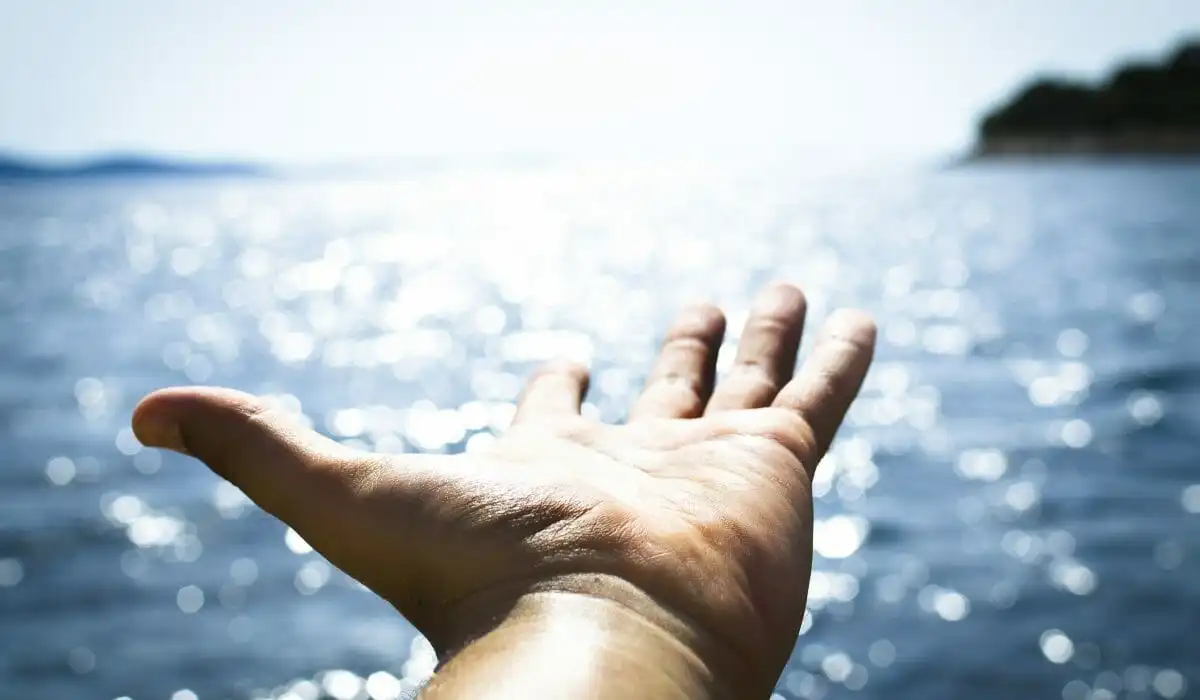 A close-up, first-person perspective of a human hand outstretched with palm open, reaching towards a sparkling, sun-drenched blue ocean. The background features blurred, glittering water bokeh and distant coastal hills under a bright sky.