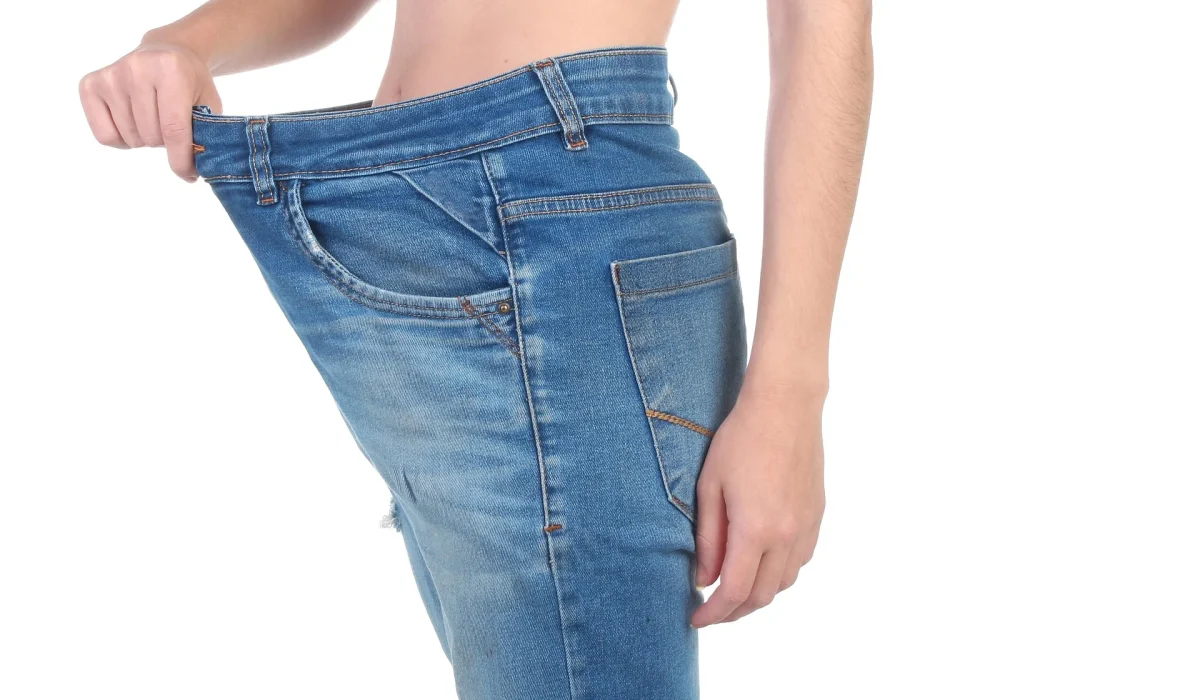 A close-up side view of a person pulling out the waistband of their blue denim jeans to show how much looser they have become around their waist.