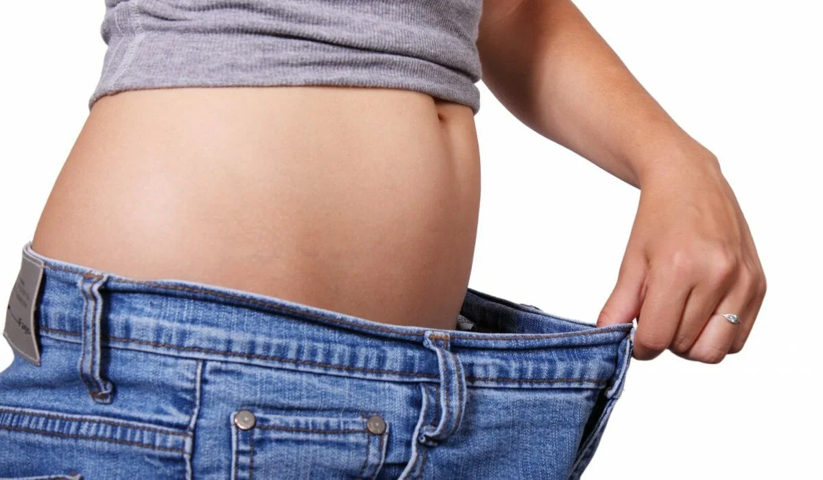 A close-up side view of a woman's flat stomach and hips. She is wearing a grey crop top and pulling the waistband of too-large blue jeans away from her waist with her hand, demonstrating significant weight loss. Isolated on a white background.
