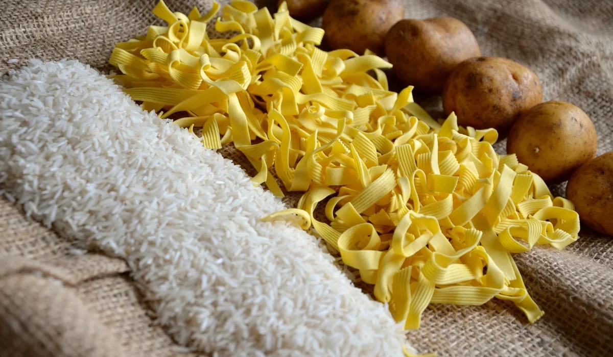 A close-up arrangement of white rice, yellow ribbon pasta, and whole brown potatoes displayed on a rustic burlap surface.