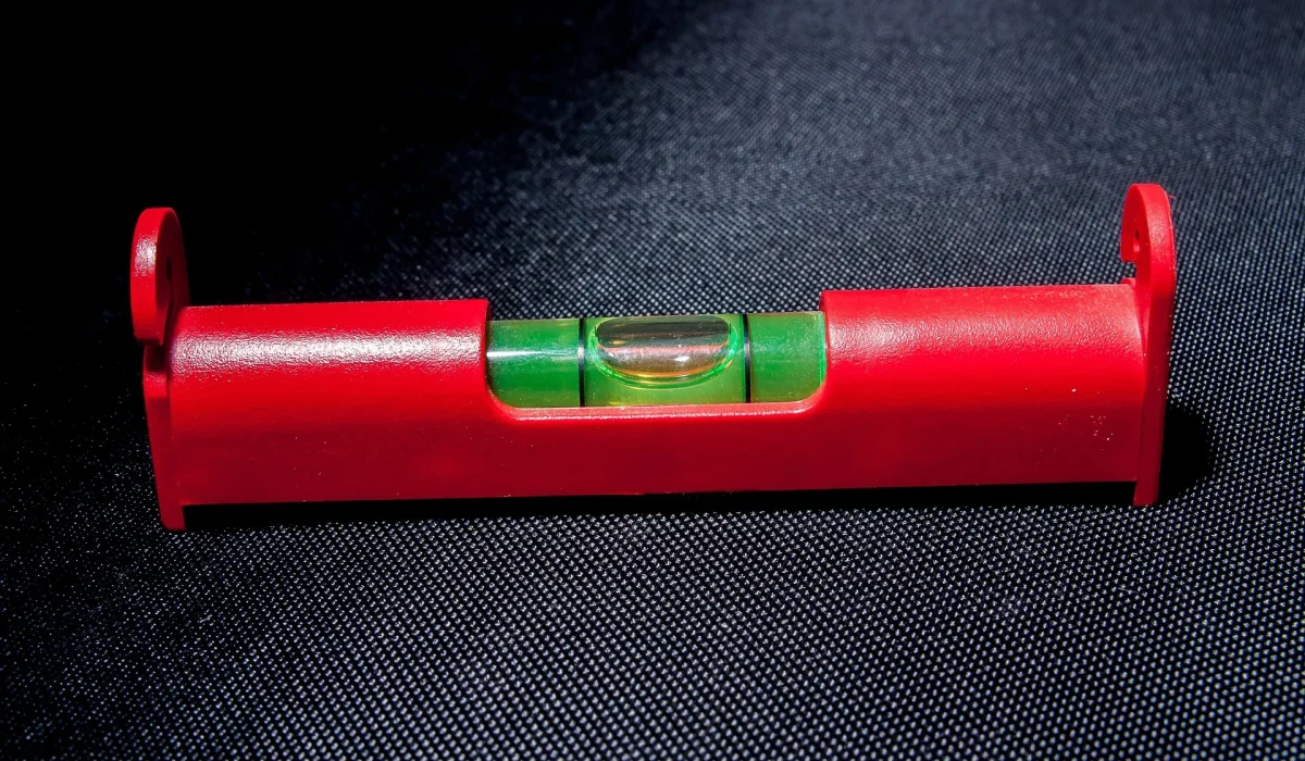 A close-up of a red spirit level tool resting on a dark, textured surface, showing the green liquid and air bubble centered between the lines.
