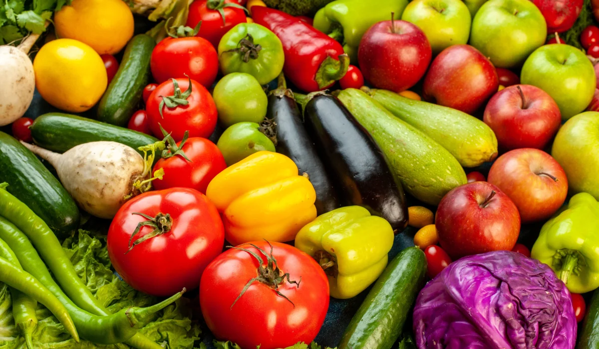 A colorful and dense top-down arrangement of various fresh vegetables and fruits, including red tomatoes, green apples, bell peppers, eggplant, cucumbers, and purple cabbage.