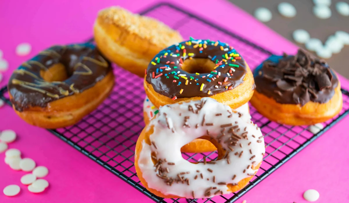 A variety of colorful donuts including chocolate glazed with sprinkles and white frosting with chocolate curls, arranged on a black wire cooling rack against a vibrant pink background.