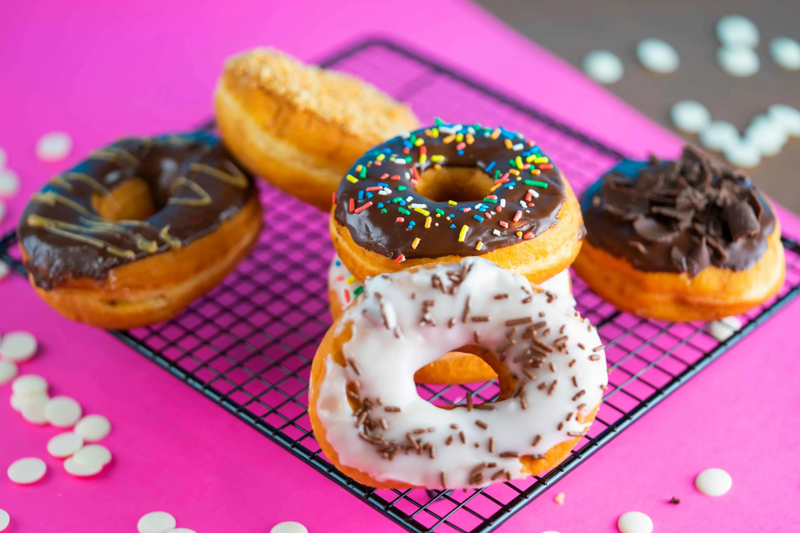 A variety of colorful donuts including chocolate glazed with sprinkles and white frosting with chocolate curls, arranged on a black wire cooling rack against a vibrant pink background.