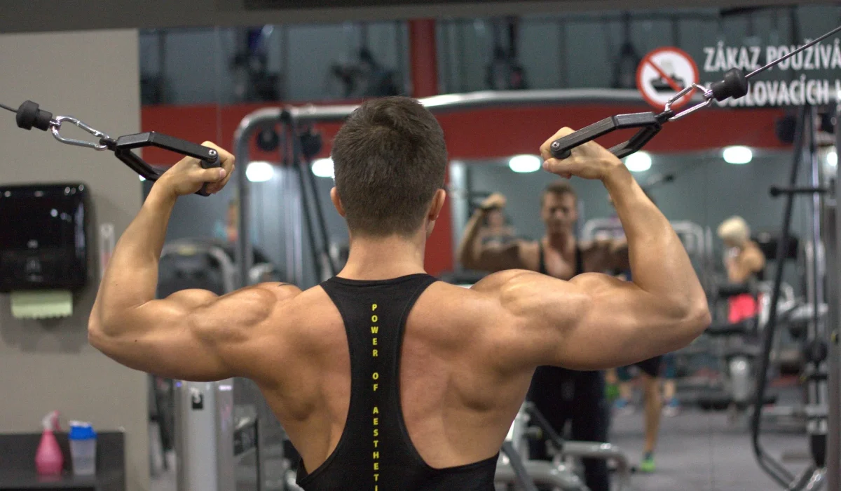 A view from behind of a muscular man performing a standing cable bicep curl, flexing his arms and back muscles in a well-lit gym.