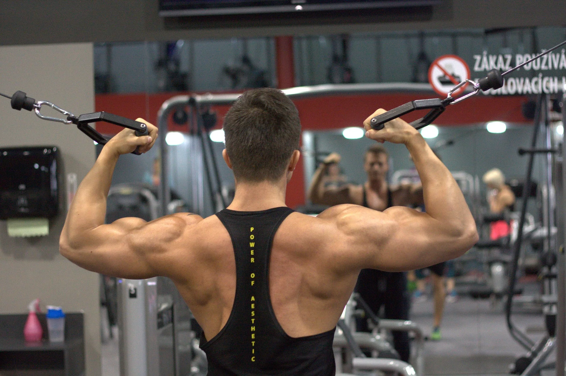 A view from behind of a muscular man performing a standing cable bicep curl, flexing his arms and back muscles in a well-lit gym.