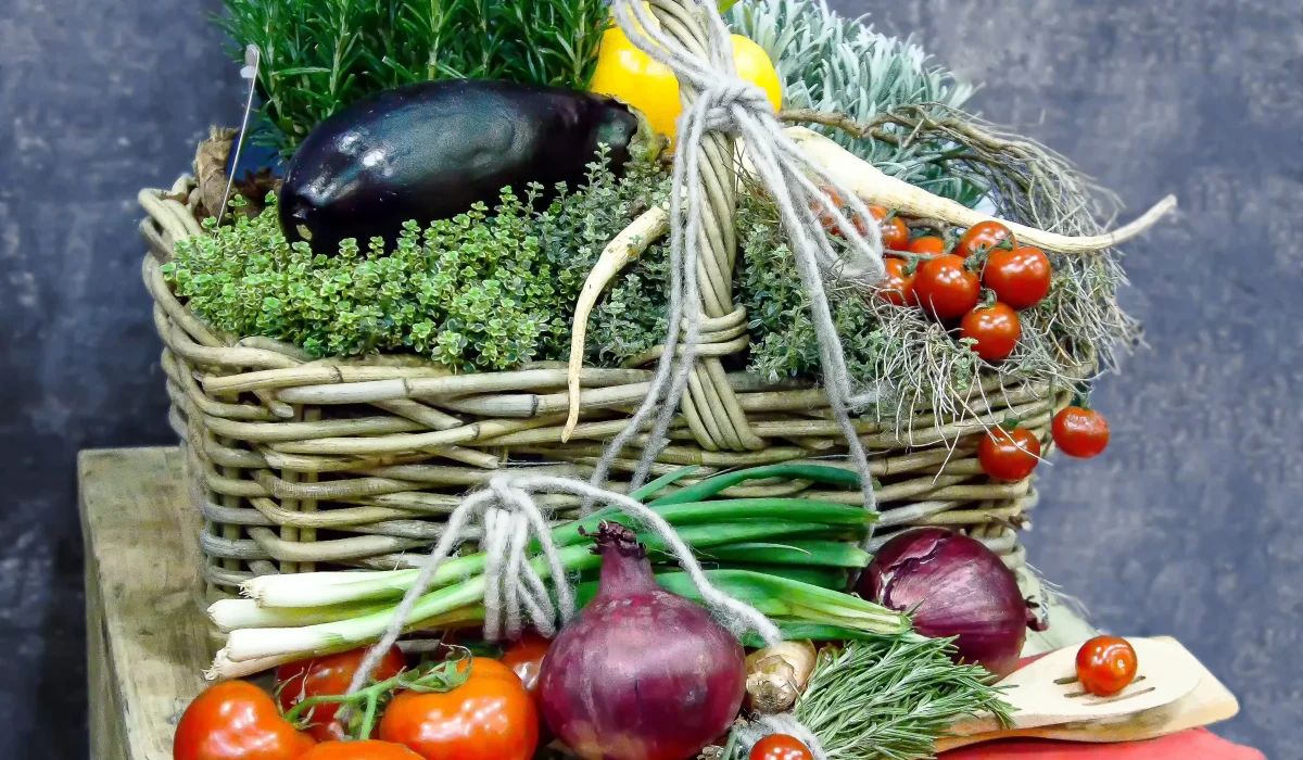 A rustic wicker basket overflowing with fresh herbs and an eggplant, surrounded by red onions, spring onions, and cherry tomatoes on a wooden surface with a pink linen accent.