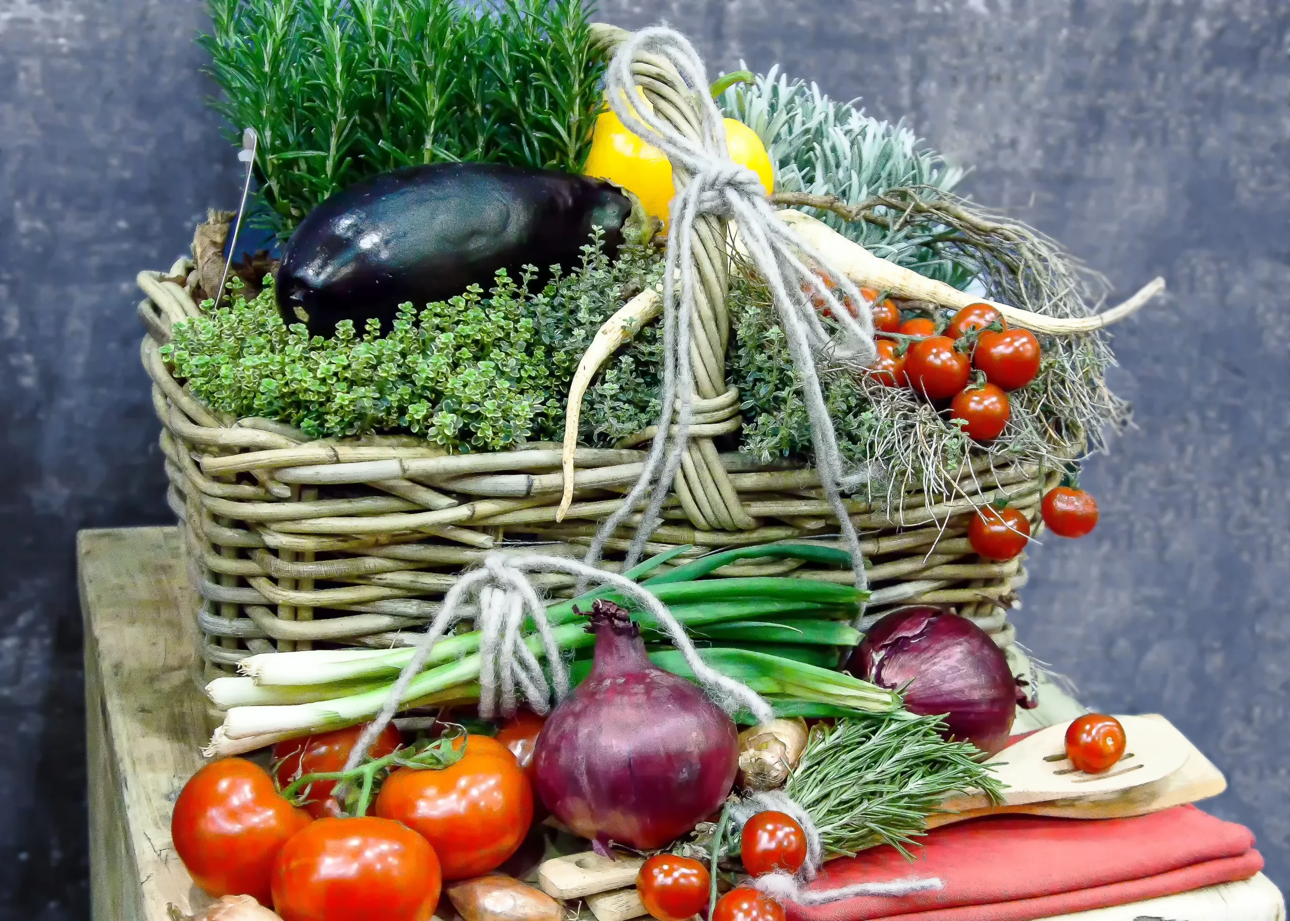 A rustic wicker basket overflowing with fresh herbs and an eggplant, surrounded by red onions, spring onions, and cherry tomatoes on a wooden surface with a pink linen accent.