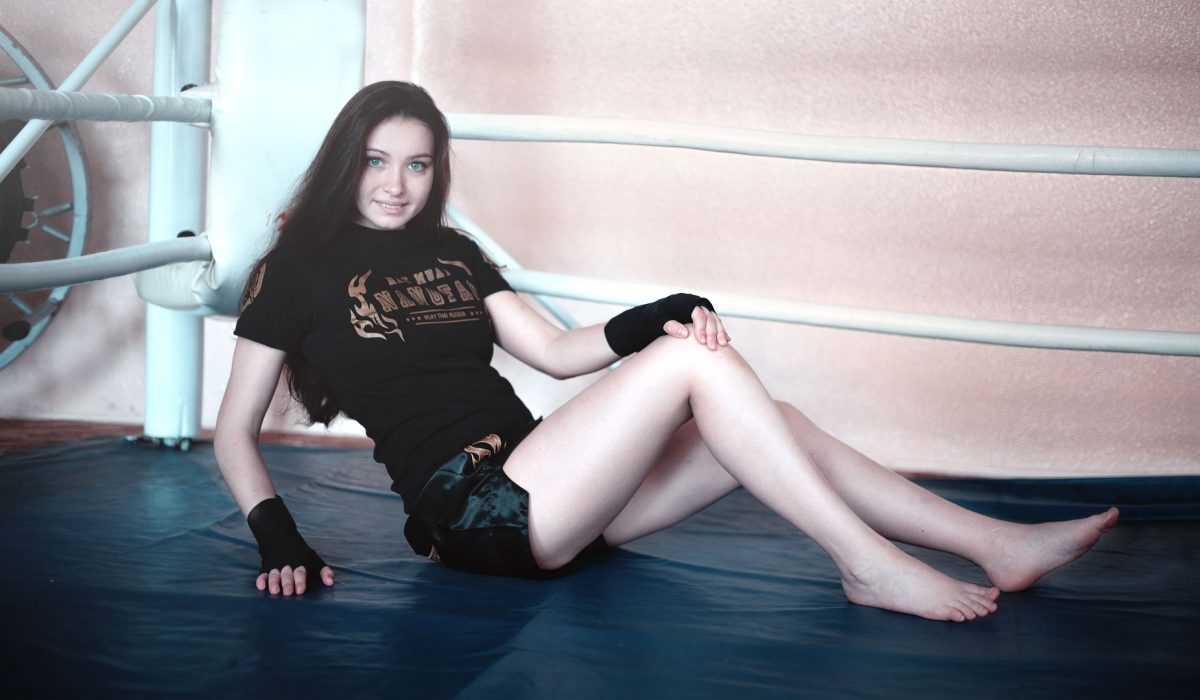 A barefoot woman in a black t-shirt and black athletic shorts with hand wraps sits and smiles in the corner of a boxing ring.