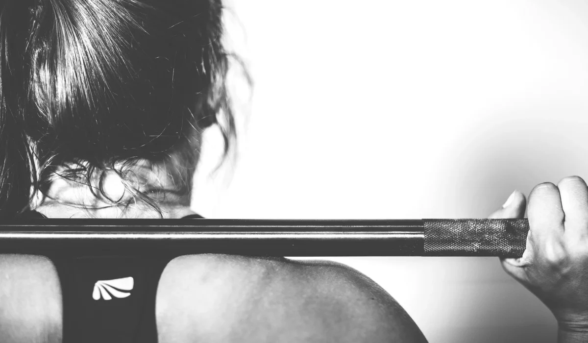 A black and white close-up from behind a woman with her hair tied up, holding a barbell across her upper back in preparation for a squat.