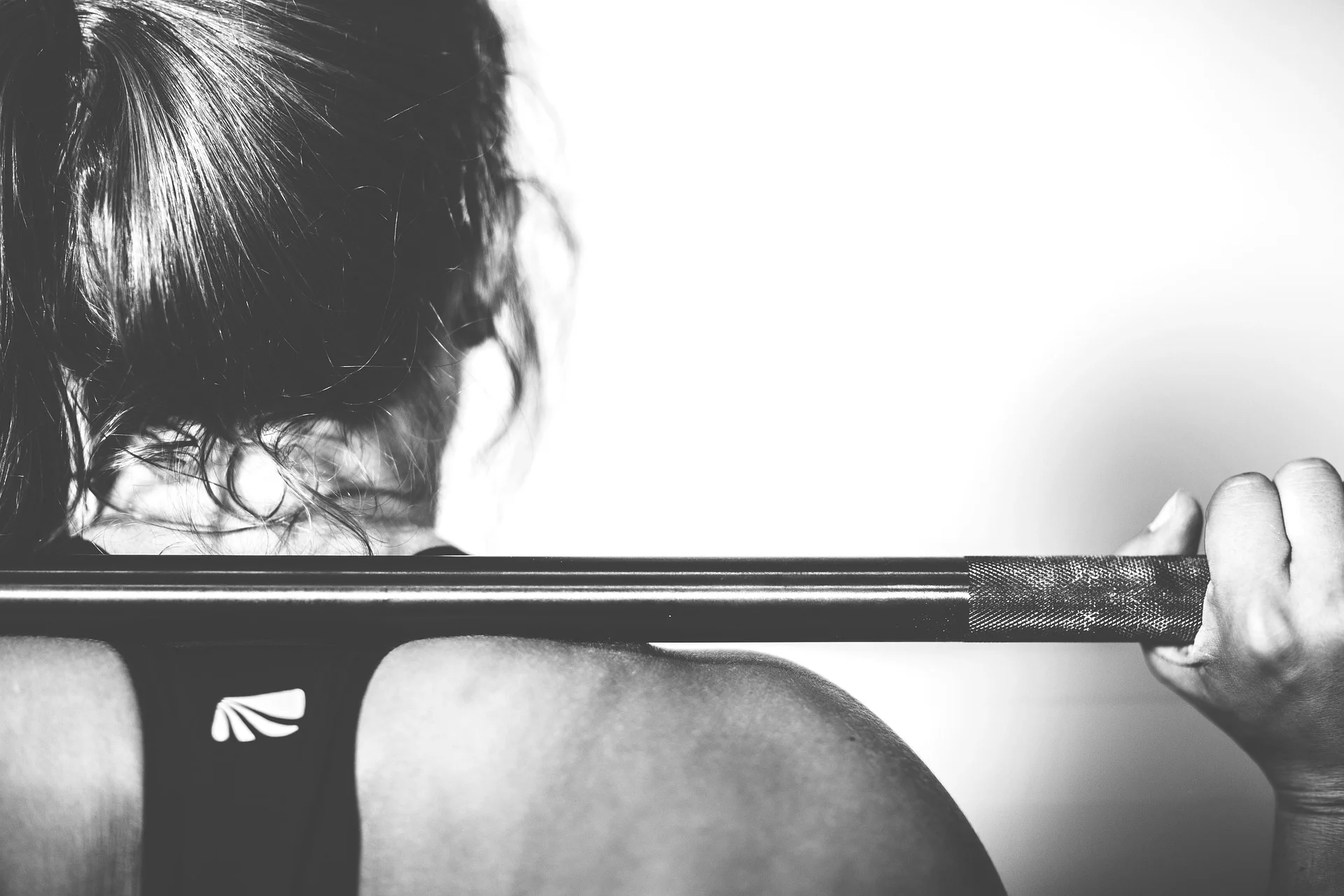 A black and white close-up from behind a woman with her hair tied up, holding a barbell across her upper back in preparation for a squat.