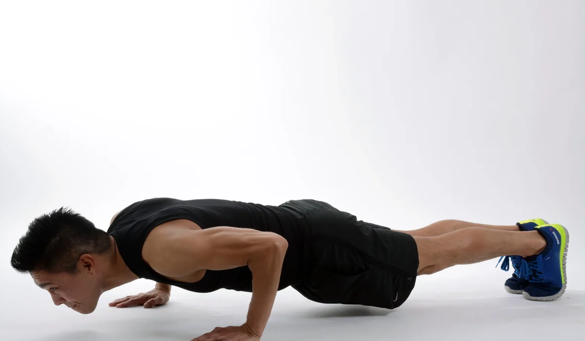 A fit man in a black tank top and shorts performing a pushup on a white background, demonstrating proper form and upper-body strength.