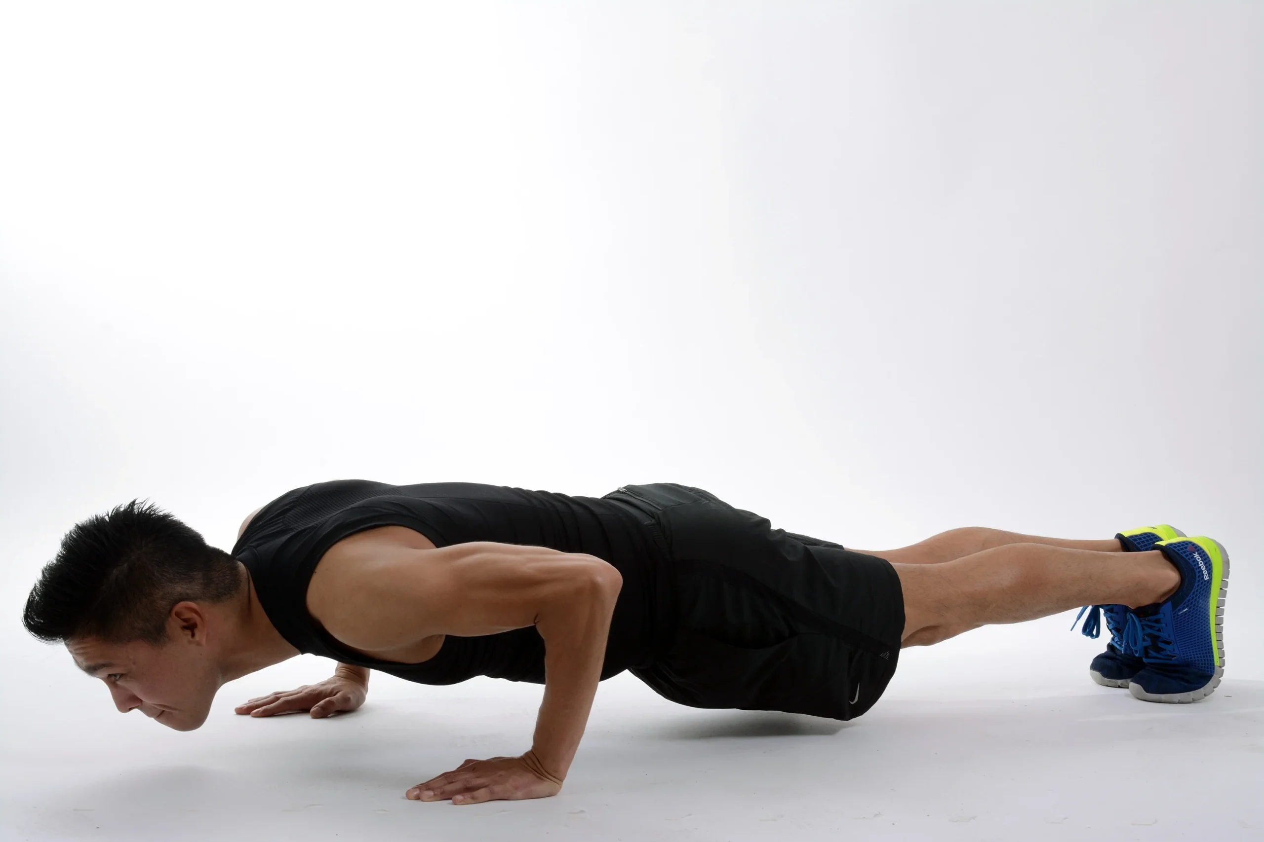A fit man in a black tank top and shorts performing a pushup on a white background, demonstrating proper form and upper-body strength.