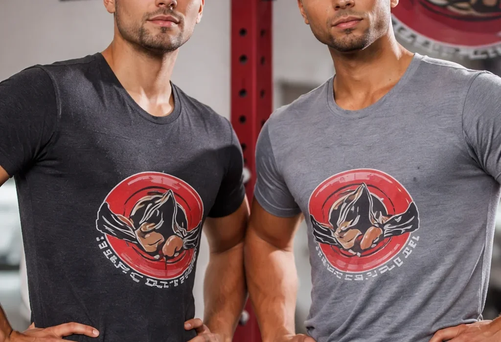 Two muscular men standing side-by-side in a gym, wearing fitted t-shirts (one charcoal, one light grey) featuring a circular red and black graphic logo on the chest.