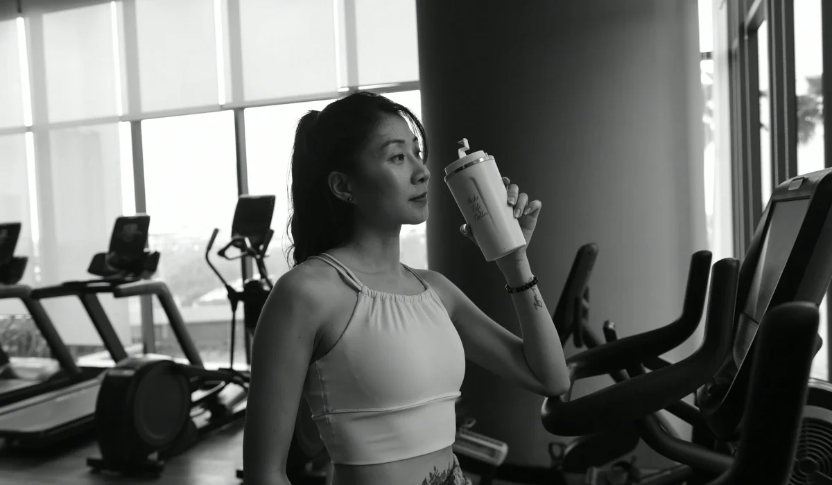 A black and white photograph of a woman in athletic wear standing in a gym, holding a shaker bottle and taking a drink, with treadmills and exercise bikes in the background.