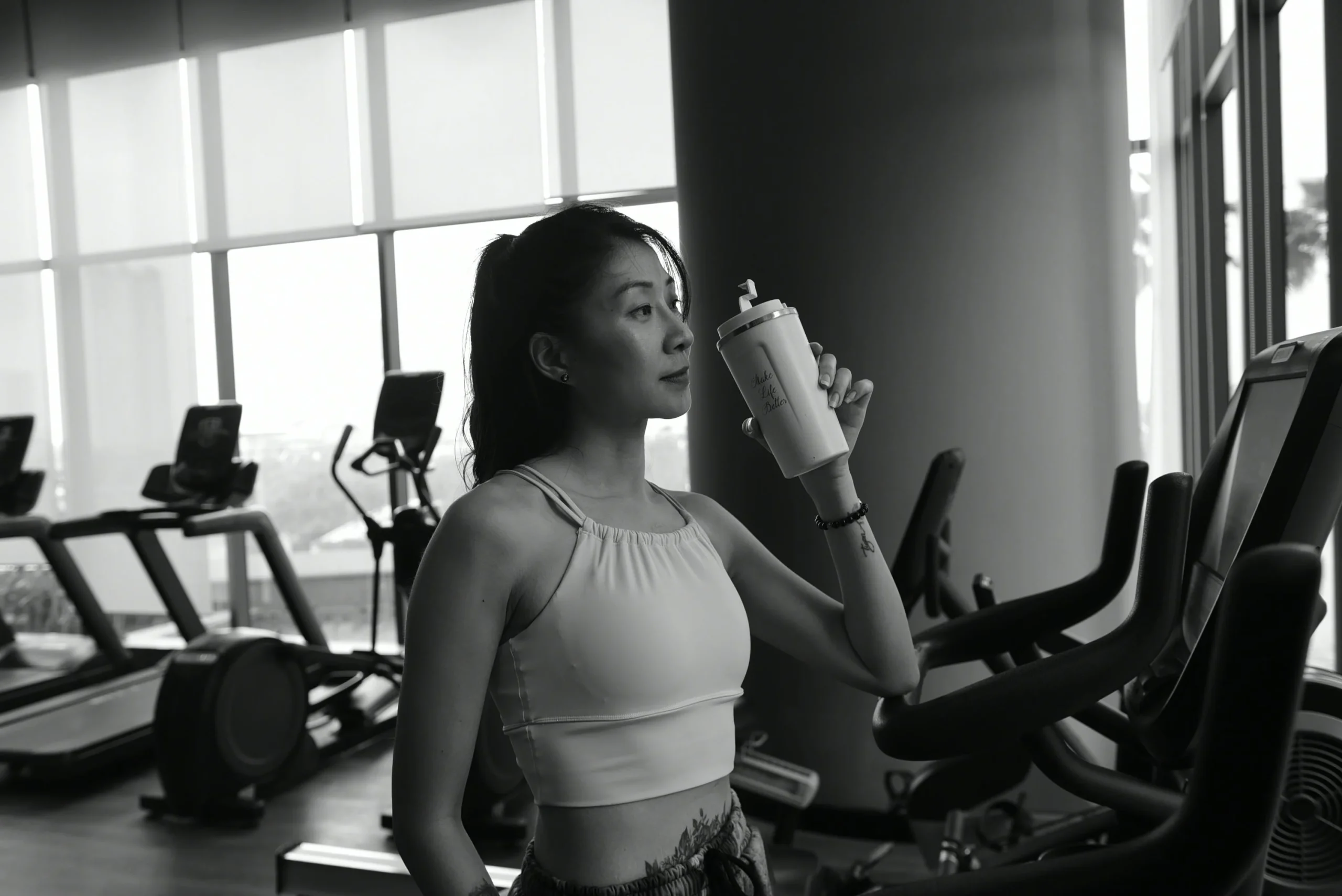 A black and white photograph of a woman in athletic wear standing in a gym, holding a shaker bottle and taking a drink, with treadmills and exercise bikes in the background.