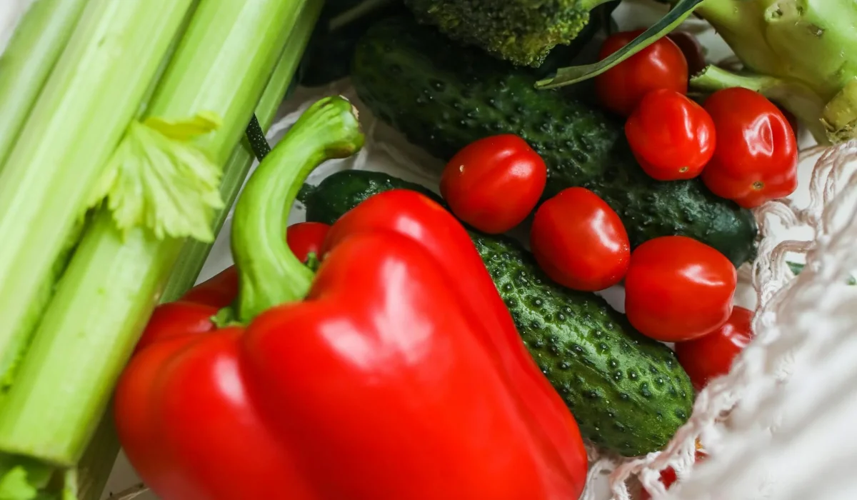 A top-down view of a white mesh reusable grocery bag filled with fresh vegetables, including a large red bell pepper, heads of broccoli, stalks of celery, cucumbers, and cherry tomatoes.