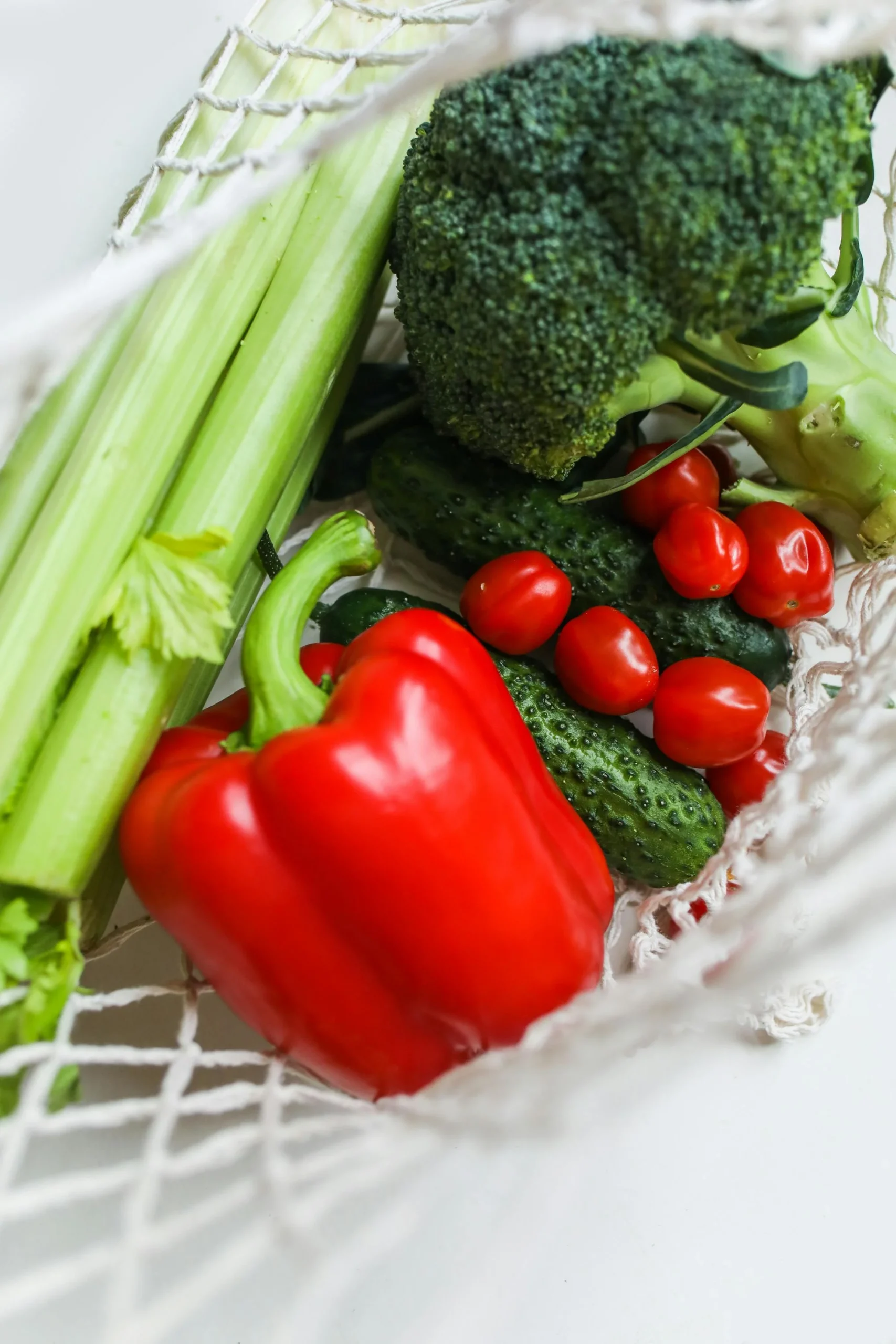 A top-down view of a white mesh reusable grocery bag filled with fresh vegetables, including a large red bell pepper, heads of broccoli, stalks of celery, cucumbers, and cherry tomatoes.