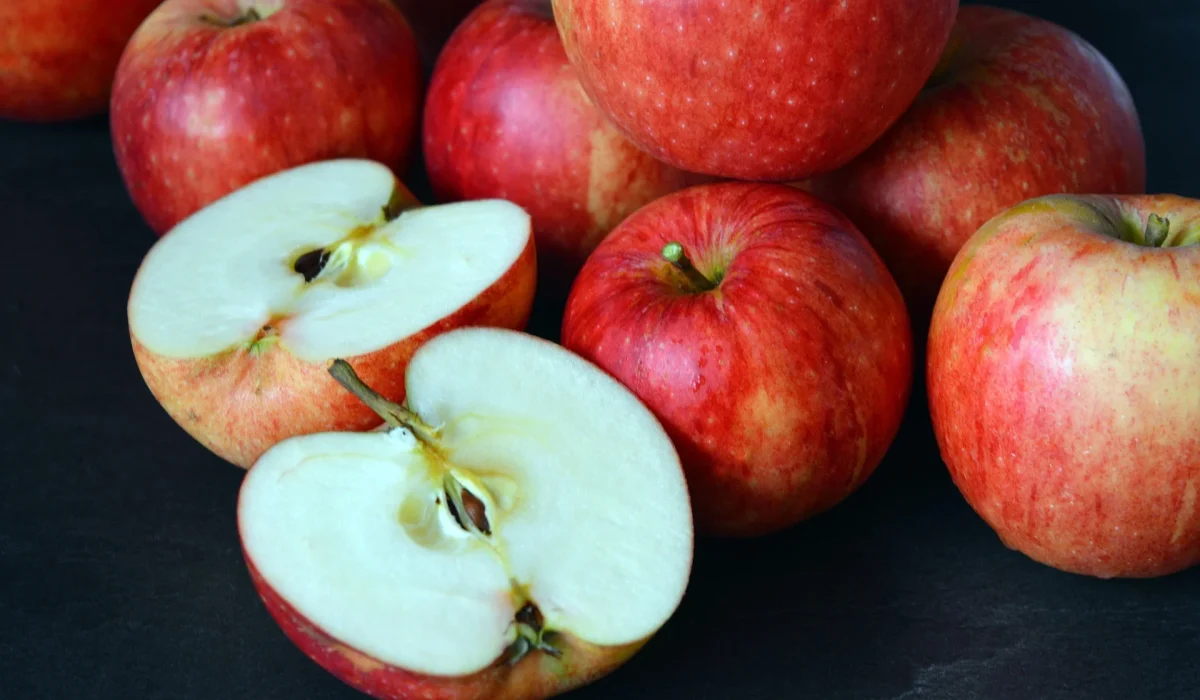 A pile of whole red gala apples with one apple sliced in half, revealing the crisp white interior and seeds, set against a dark background.