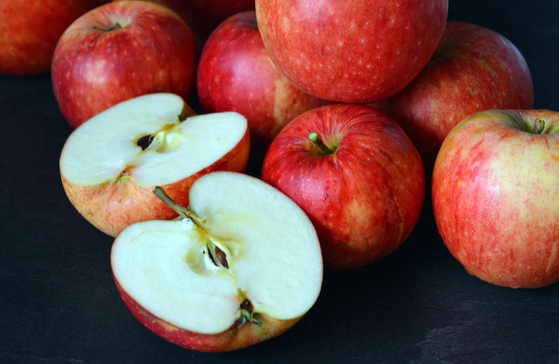 A pile of whole red gala apples with one apple sliced in half, revealing the crisp white interior and seeds, set against a dark background.