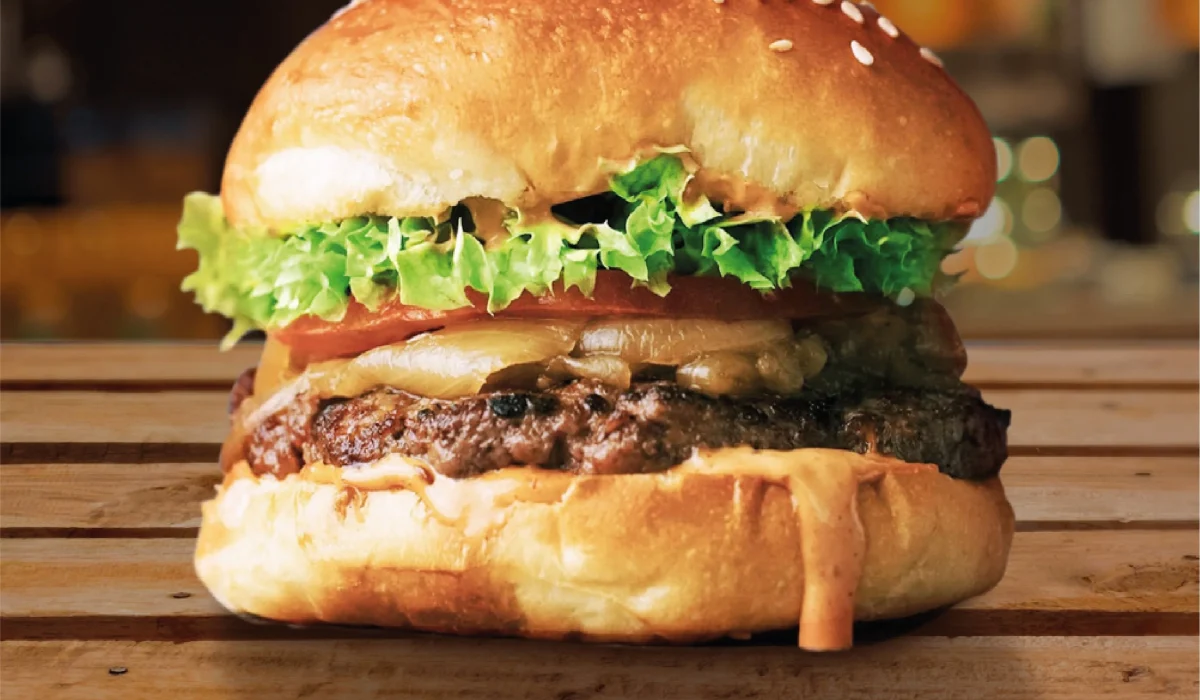 A close-up side view of a juicy beef burger on a toasted sesame seed bun, layered with fresh lettuce, tomato, melted cheese, and a signature sauce on a wooden surface.