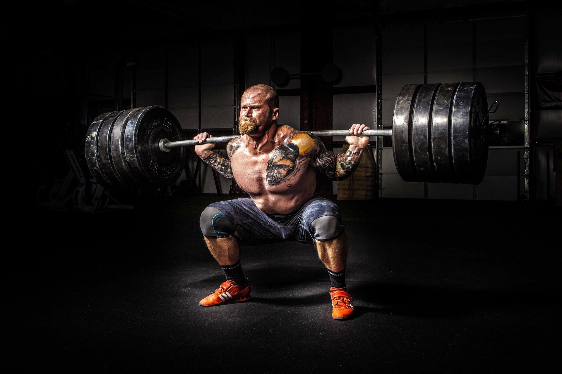 A muscular, tattooed man performing a heavy back squat with a loaded barbell in a dark gym under dramatic spotlighting.