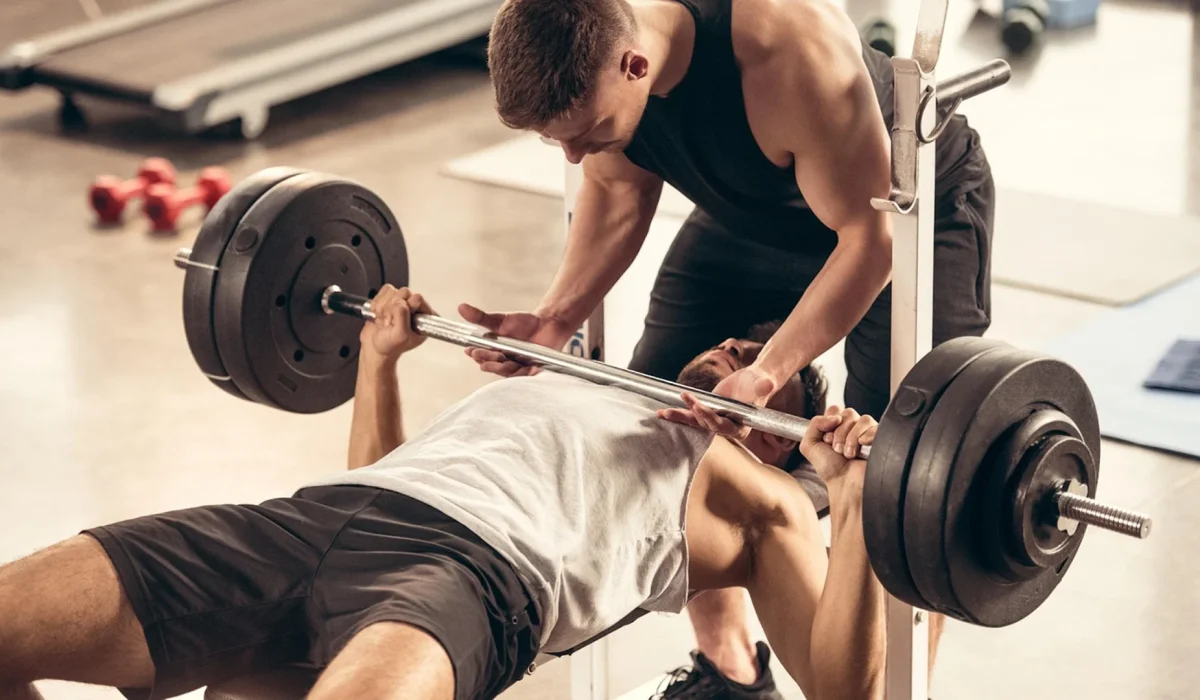 A man performing a heavy barbell bench press on a weight bench while being closely spotted by another man in a brightly lit, professional gym setting.