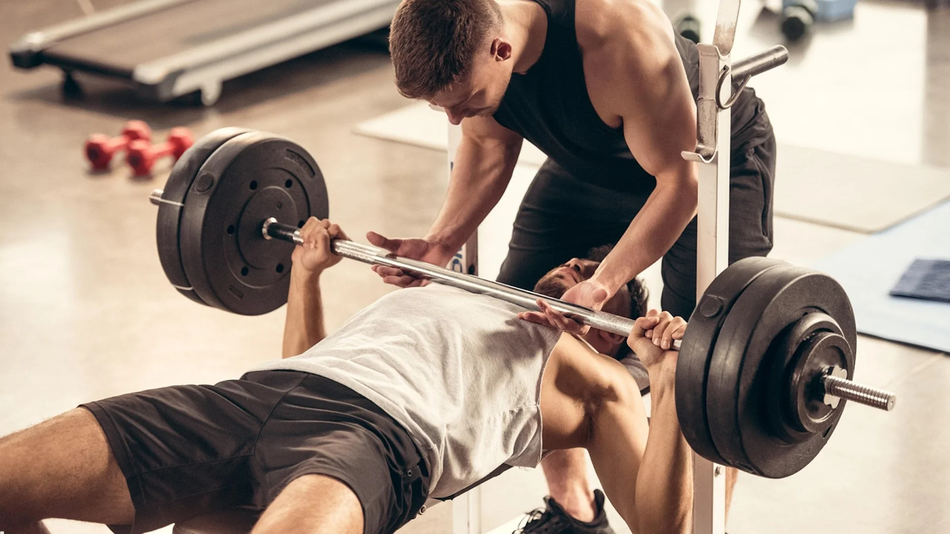 A man performing a heavy barbell bench press on a weight bench while being closely spotted by another man in a brightly lit, professional gym setting.