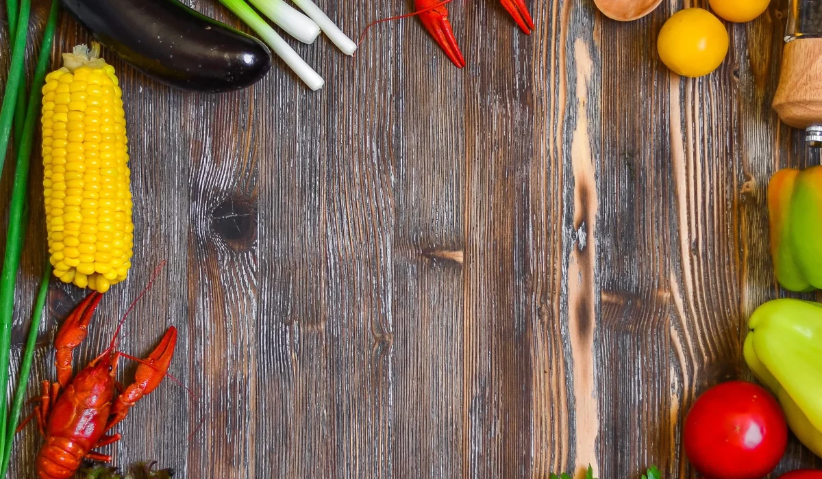 A top-down view of various fresh ingredients creating a border on a rustic wooden table, including shrimp, a pork chop, fresh greens, tomatoes, corn, and a whole coconut.