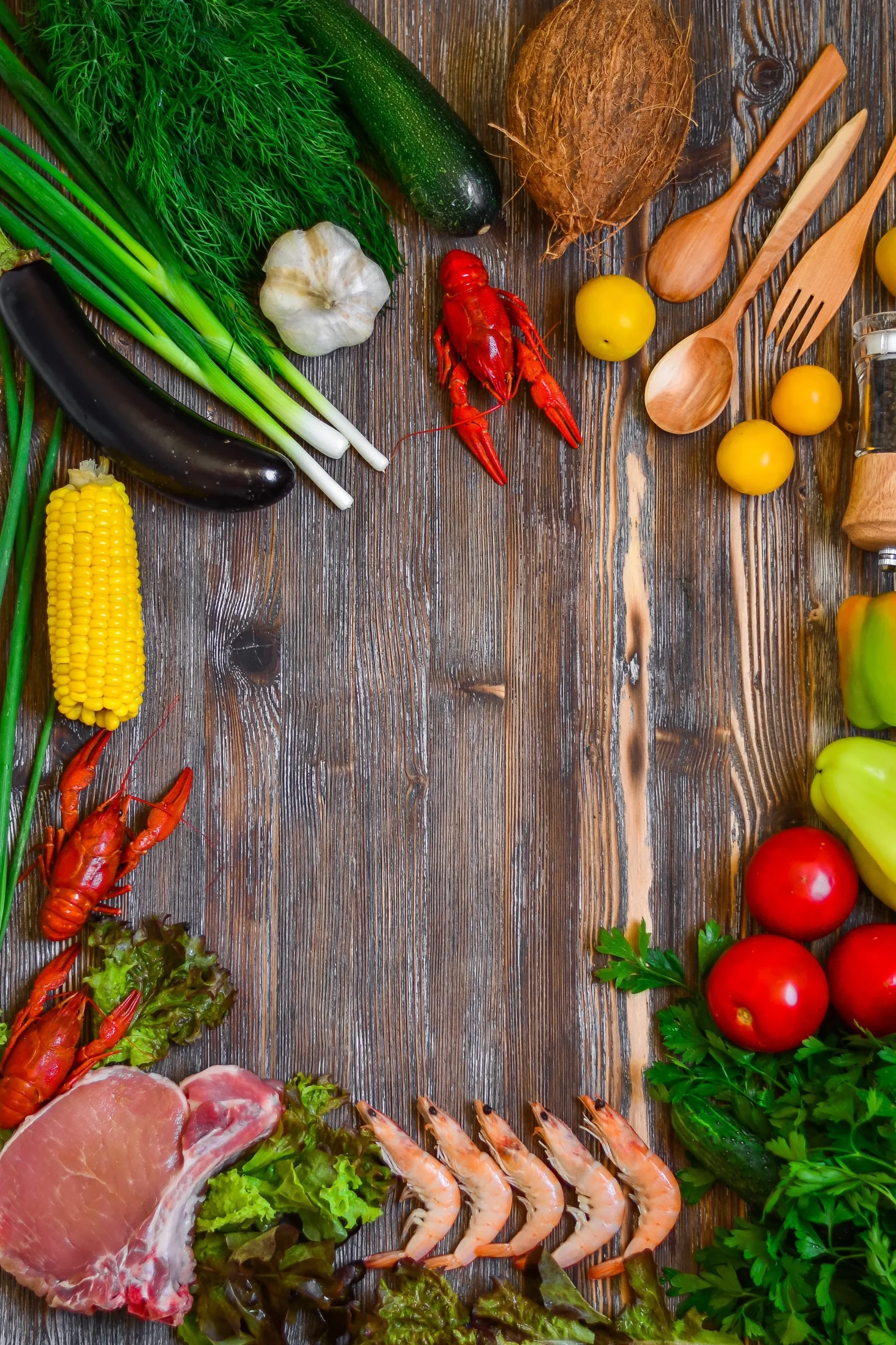 A top-down view of various fresh ingredients creating a border on a rustic wooden table, including shrimp, a pork chop, fresh greens, tomatoes, corn, and a whole coconut.