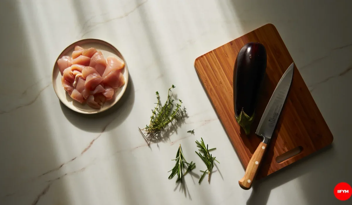 A top-down view of fresh meal prep ingredients on a marble surface including raw diced chicken breast on a small plate, an eggplant on a wooden cutting board with a chef's knife, and fresh sprigs of rosemary and thyme, featuring the IIFYM logo.