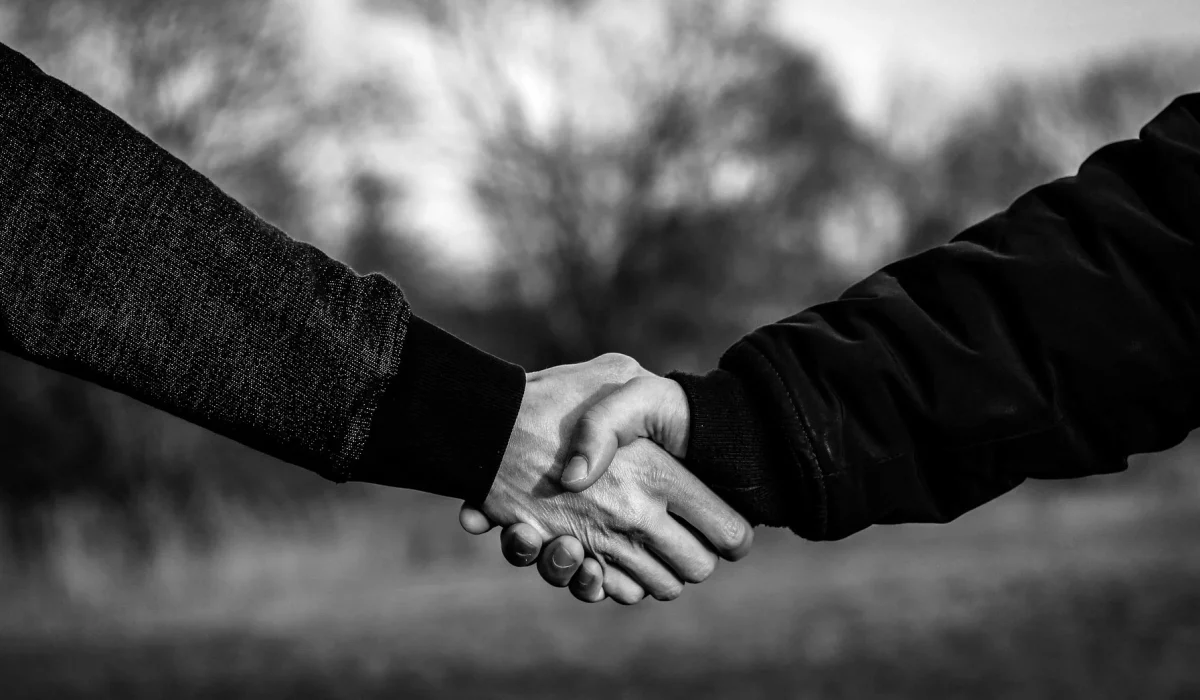 A close-up, black and white photograph of two individuals in jackets shaking hands outdoors against a soft, blurred natural background.