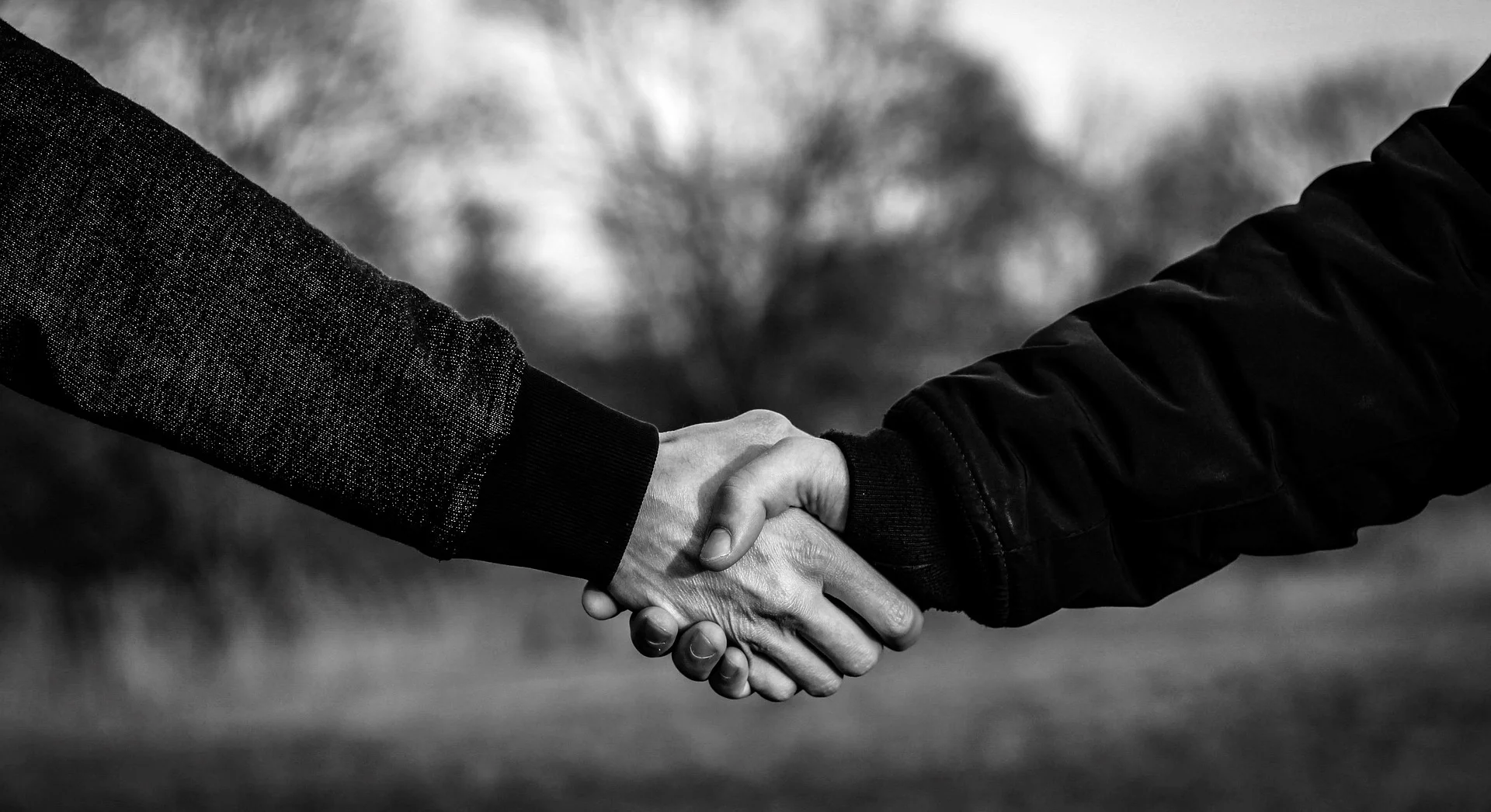 A close-up, black and white photograph of two individuals in jackets shaking hands outdoors against a soft, blurred natural background.