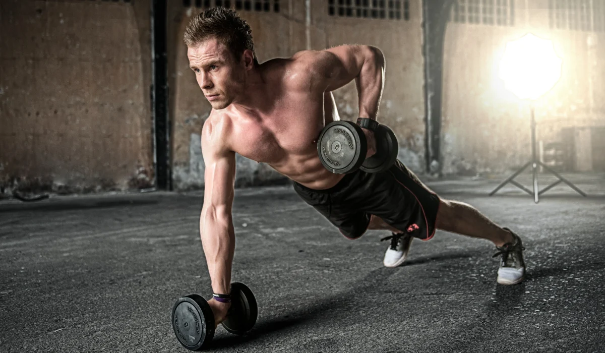 A shirtless man in an industrial-style gym performing a renegade row, balancing in a plank position on dumbbells while rowing one weight toward his hip.