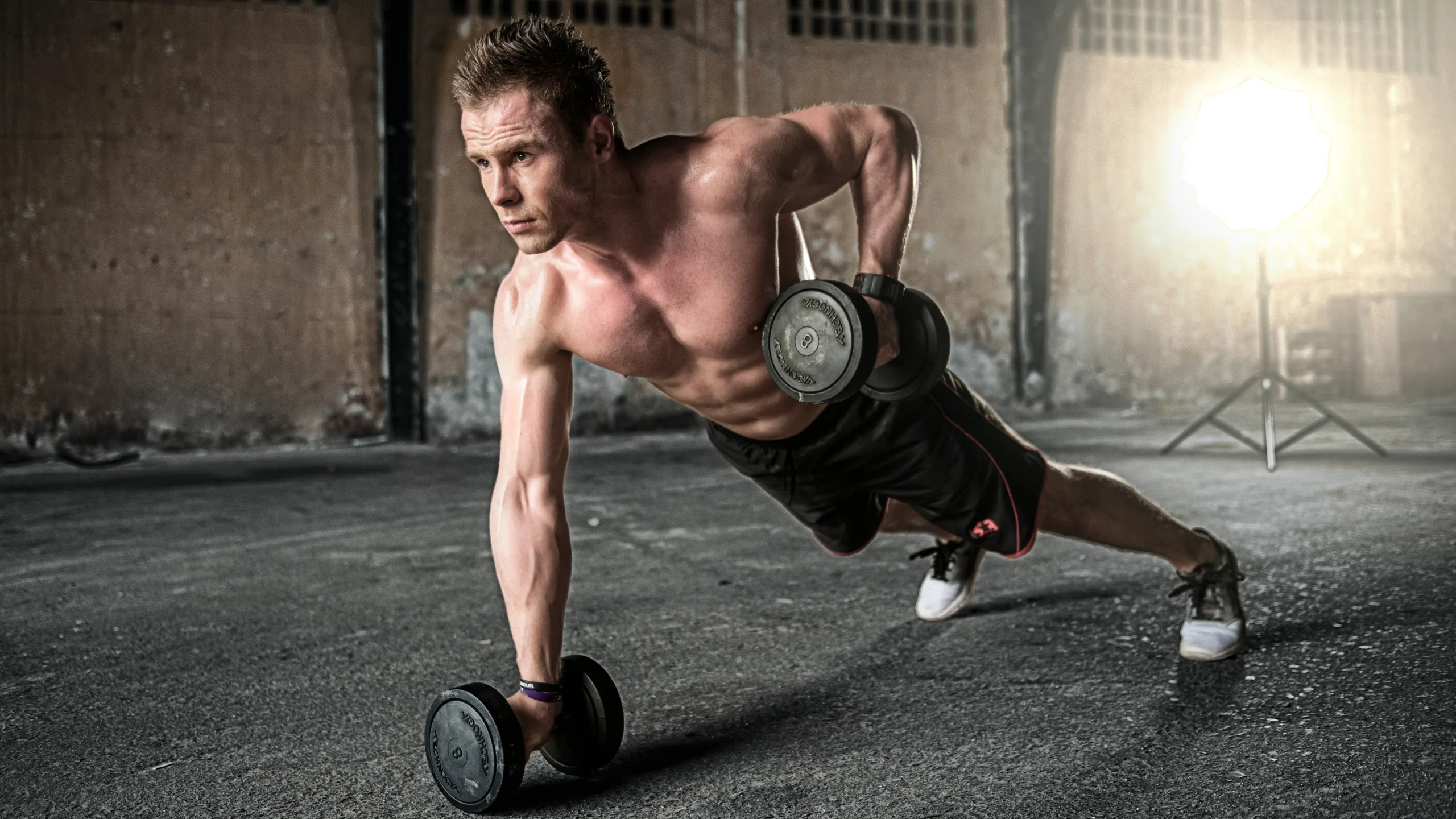 A shirtless man in an industrial-style gym performing a renegade row, balancing in a plank position on dumbbells while rowing one weight toward his hip.
