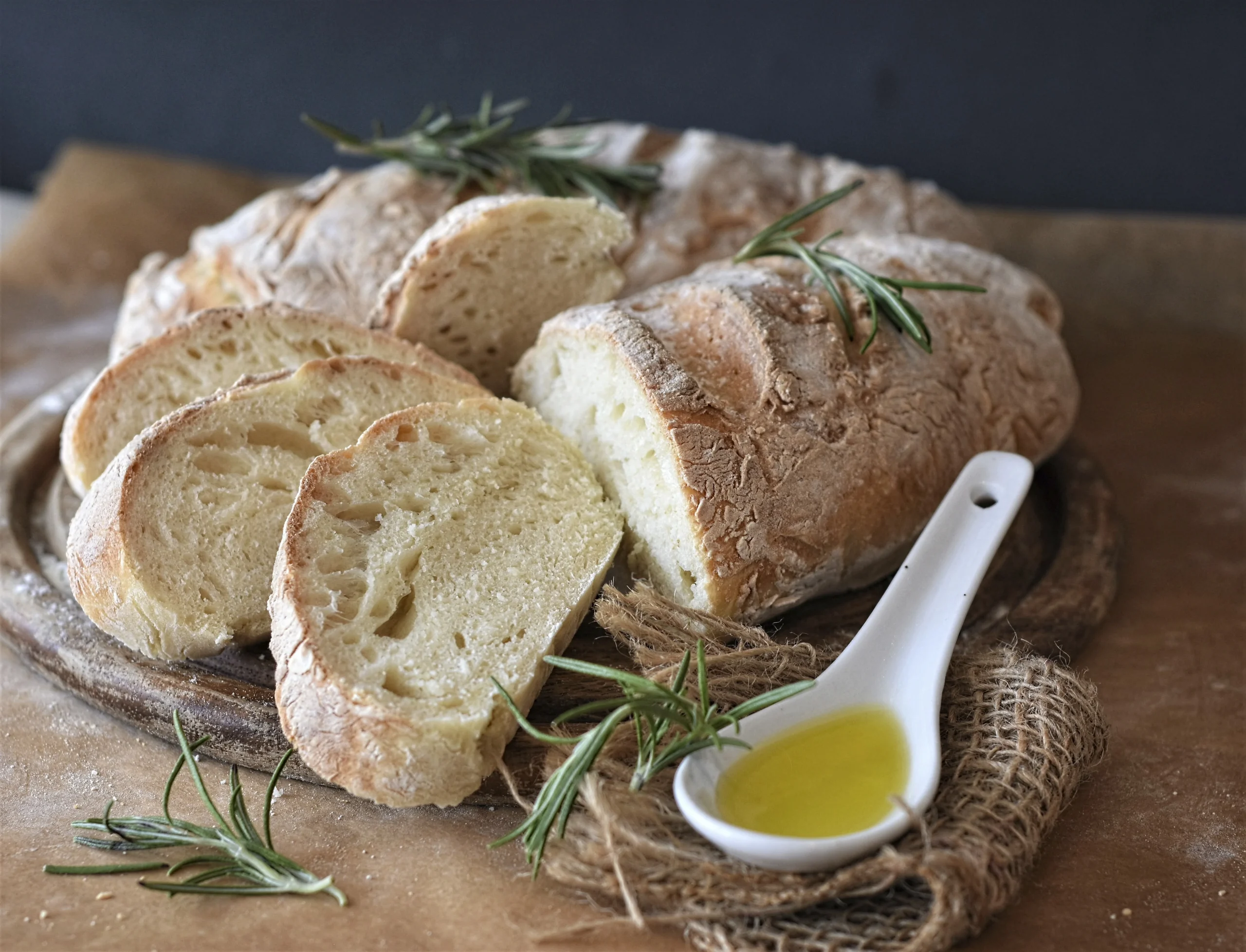 Slices of fresh, crusty artisan bread topped with rosemary sprigs on a wooden board, served with a small white spoon of golden olive oil.