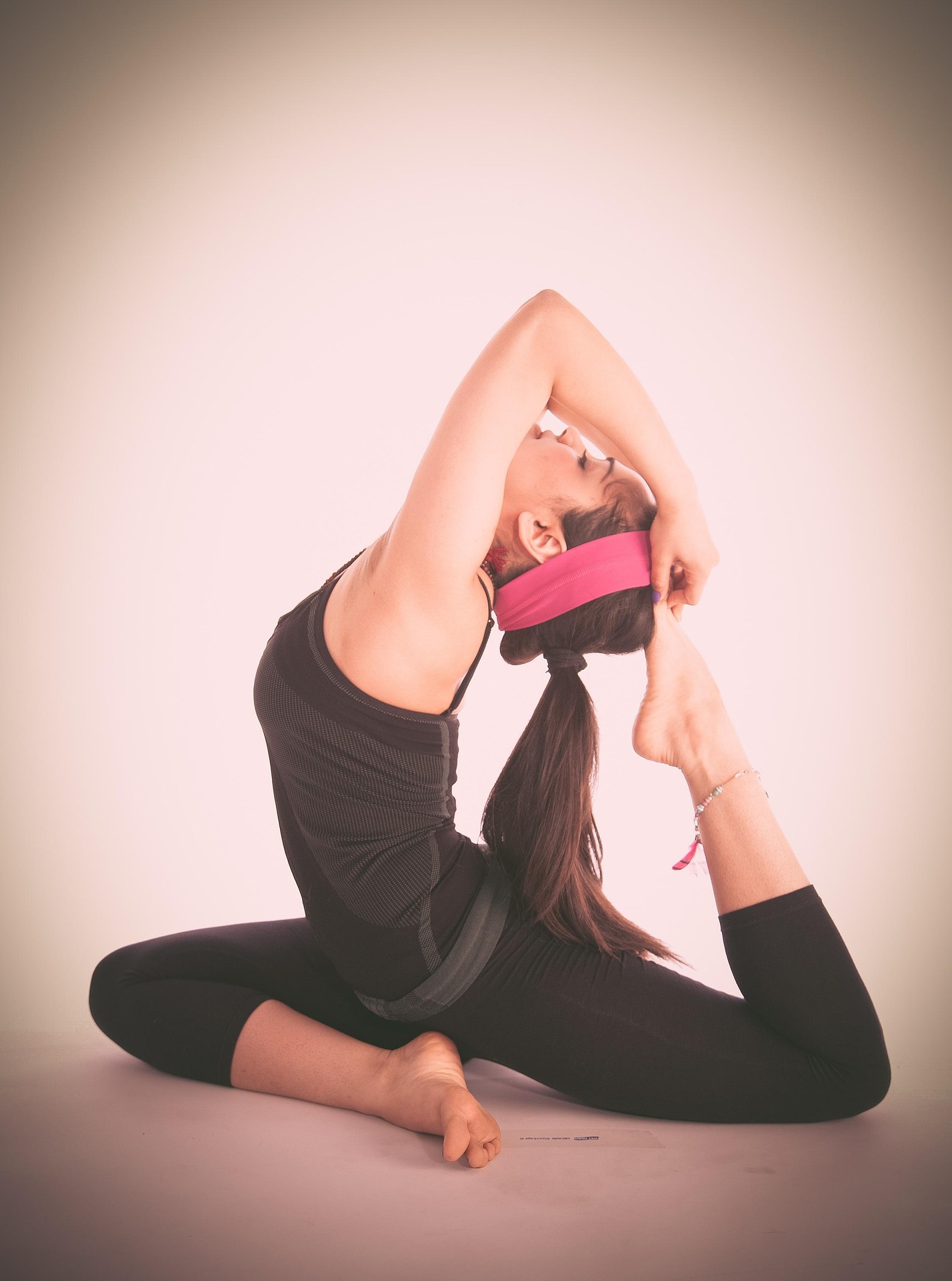 A woman with a long ponytail and pink headband performing the One-Legged King Pigeon yoga pose against a plain light background.