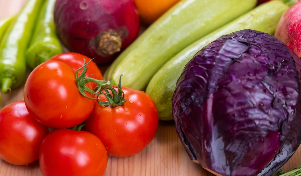A colorful assortment of fresh organic vegetables on a light wooden cutting board. Includes red tomatoes on the vine, a whole purple cabbage, green zucchini, green peppers, and a purple turnip. The vegetables show natural texture and water droplets.