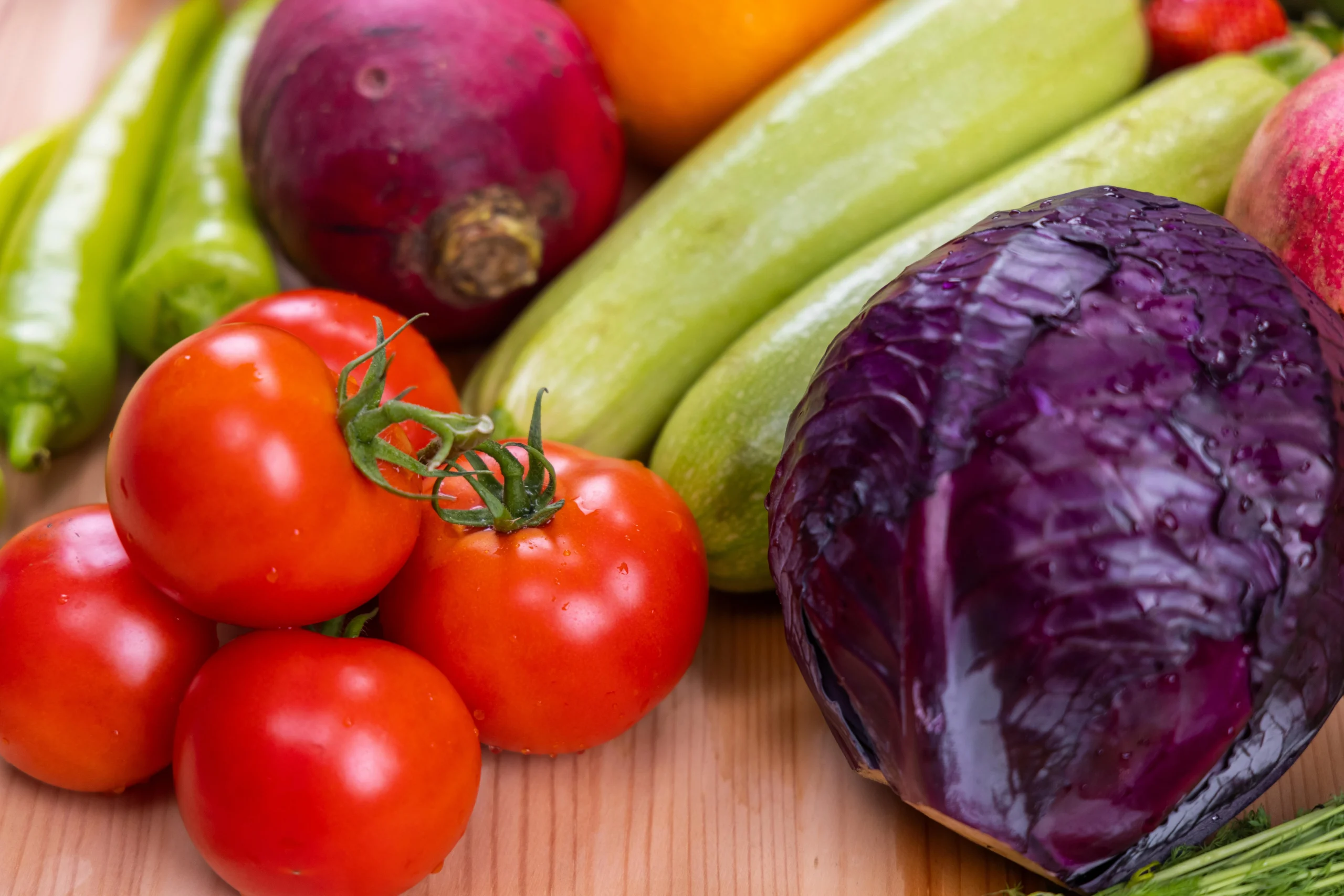 A colorful assortment of fresh organic vegetables on a light wooden cutting board. Includes red tomatoes on the vine, a whole purple cabbage, green zucchini, green peppers, and a purple turnip. The vegetables show natural texture and water droplets.