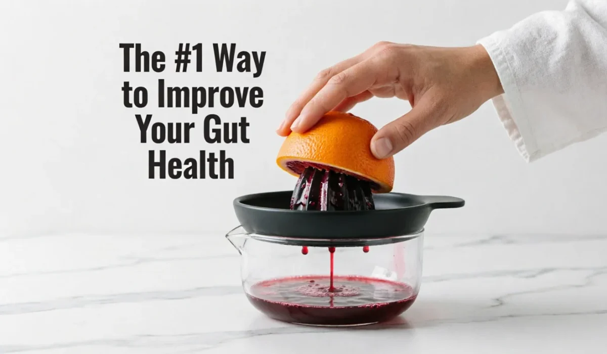 A close-up of a person's hand juicing a blood orange using a manual black and glass juicer on a white marble surface. Red juice is dripping into the glass container. Text on the left reads "The #1 Way to Improve Your Gut Health."
