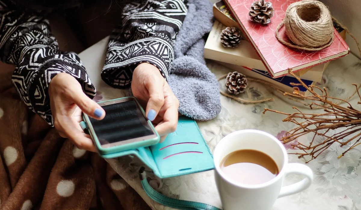 A top-down view of a person wearing a patterned sweater holding a smartphone in a teal case, with a white mug of coffee, books, and pine cones resting on a table nearby.