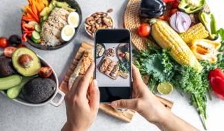 A top-down view of hands holding a smartphone to photograph a variety of healthy foods, including an egg and rice bowl, avocados, corn, and fresh greens spread across a table.