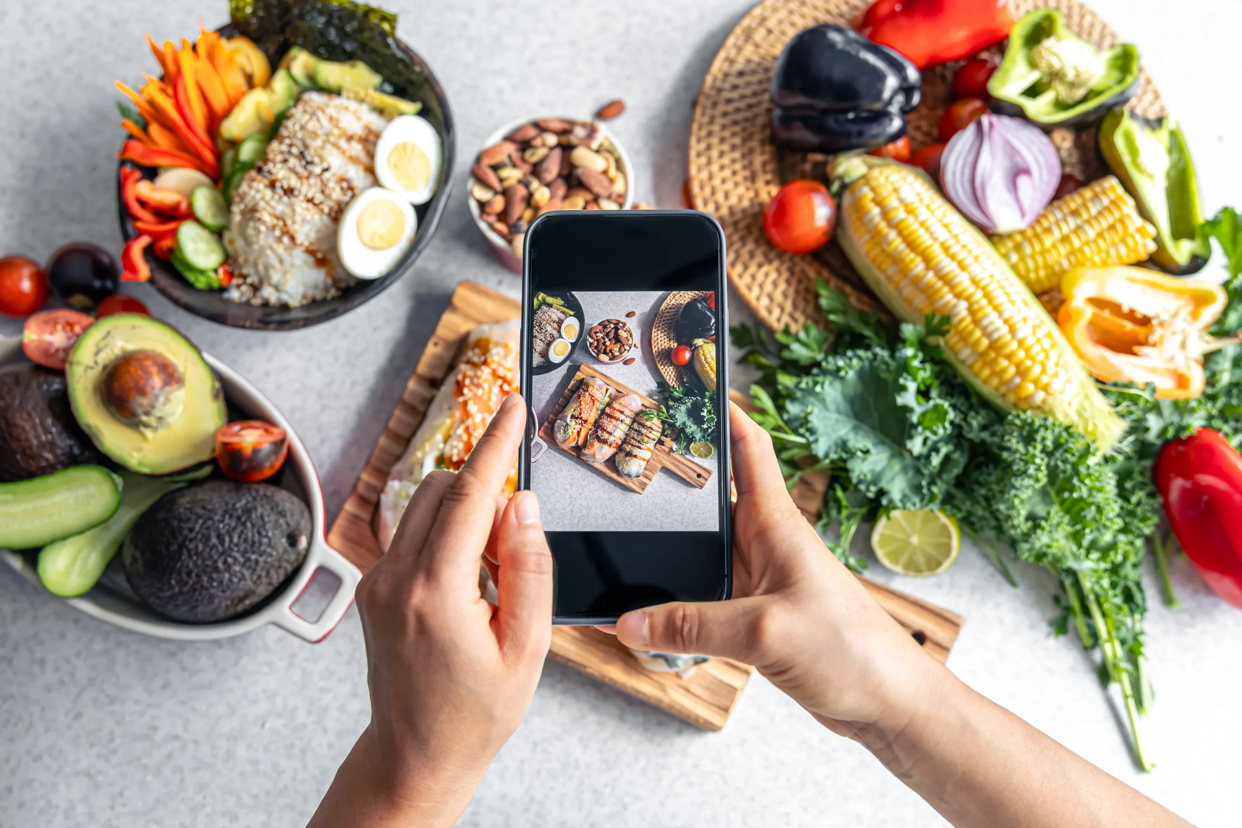 A top-down view of hands holding a smartphone to photograph a variety of healthy foods, including an egg and rice bowl, avocados, corn, and fresh greens spread across a table.